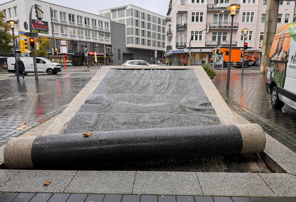 Im Vordergrund befindet sich ein großes, rechteckiges Wasserbecken mit einer leicht geneigten, glatten Steinfläche, über die Wasser in dünnem Film nach unten fließt. Am unteren Rand rollt sich ein massiver, dunkler Steinbalken leicht nach oben. Im Hintergrund stehen mehrstöckige Gebäude mit zahlreichen Fenstern und Balkonen, einige mit Geschäften im Erdgeschoss. Die Straße und der Gehweg sind nass, was auf Regenwetter hindeutet. Mehrere Fahrzeuge, darunter ein weißer Transporter und ein Auto, sind zu sehen. Die Szene wirkt urban und belebt.