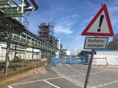 Foto Chempark Dormagen. Schild mit Ausrufezeichen, darunter: Radfahrer Absteigen. Industrie und Straße im Hintergurnd, ein verschlossenes Tor.