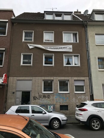 Foto von einem Haus. Im Erdgeschoss Graffiti an der Wand. Über dem ersten und unter dem zweiten Stockwerk hängt ein Banner, durch den Wind leicht verdreht, mit der Aufschrift: WWW.CORONA-AUSSCHUSS.de
