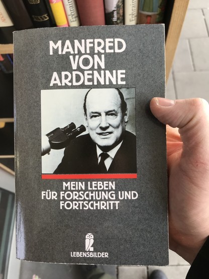 Buch "Manfred von Ardenne - Mein Leben für Forschung und Fortschritt" aus dem Verlag "Lebensbilder" mit einem Foto des Autors auf dem Cover. Er trägt Anzug, weißes Hemd und Krawatte, schaut direkt in die Kamera und hat ein Mikroskop neben sich. Im Hintergrund ein öffentlicher Bücherschrank. Das Buch wird von einer Hand gehalten, ein rechter Daumen fixiert das Buch um es mit der linken Hand (nicht im Bild) fotografieren zu können.